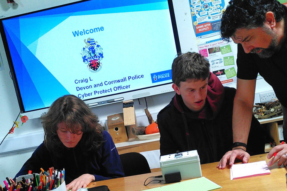 A police cyber safety talk in a classroom, with a screen displaying "Welcome, Craig L, Devon and Cornwall Police, Cyber Protect Officer." A student sits at a desk while an instructor leans in to point at his notebook.