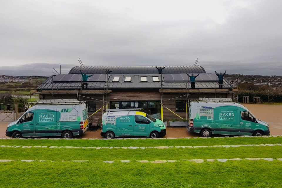 Three green Naked Solar branded vans parked outside a building with a curved roof, while installers stand on scaffolding on the roof with arms raised, having just fitted solar panels.
