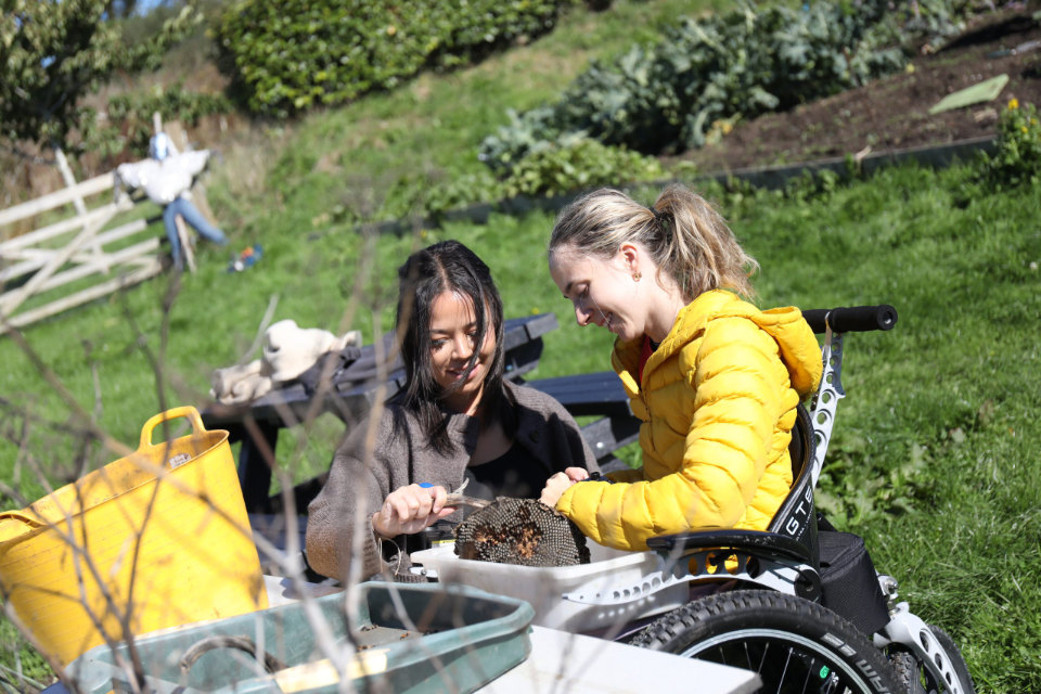 Two women sit at an outdoor table working together on a craft project involving a large dried sunflower head, with garden beds and a scarecrow visible behind them.