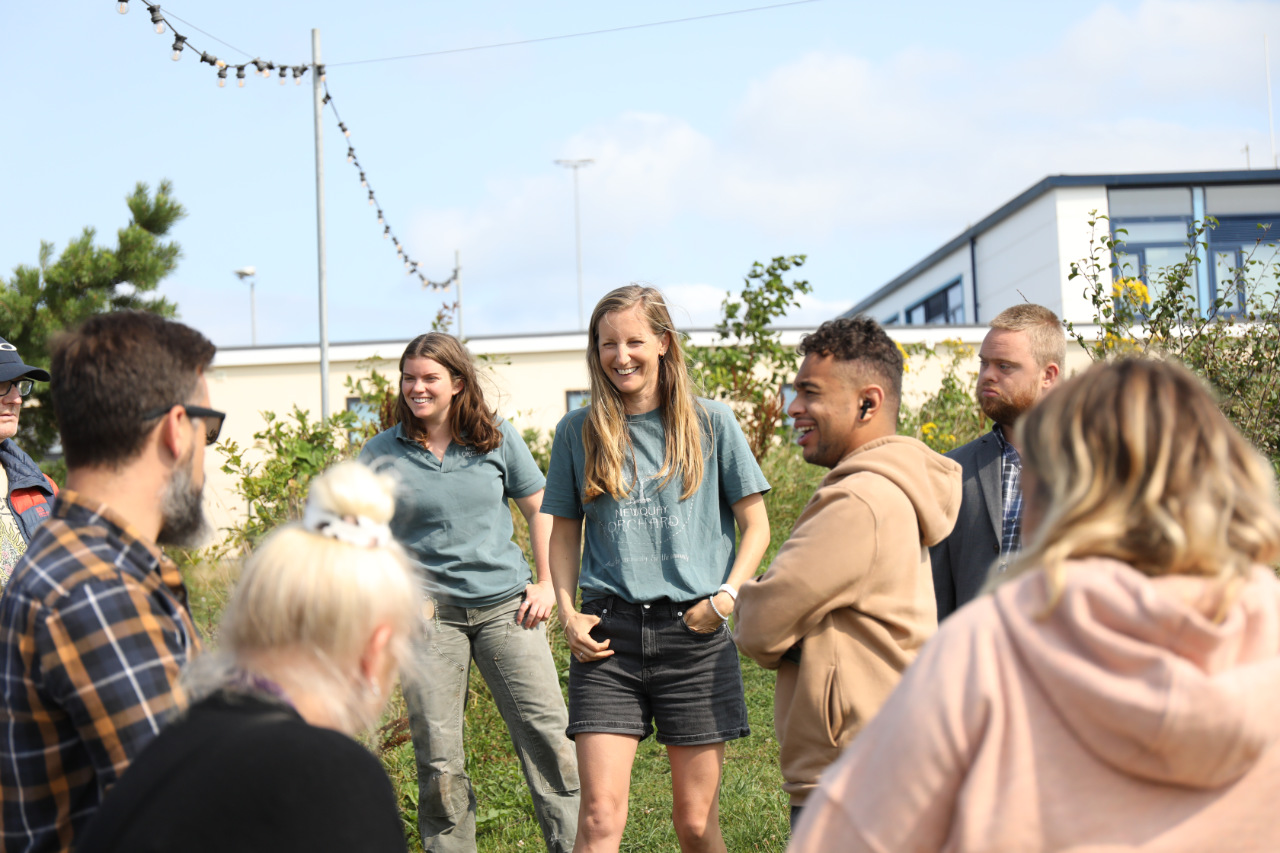 A group of people standing outdoors in a grassy area, gathered in a loose circle and smiling/laughing during a community garden gathering.