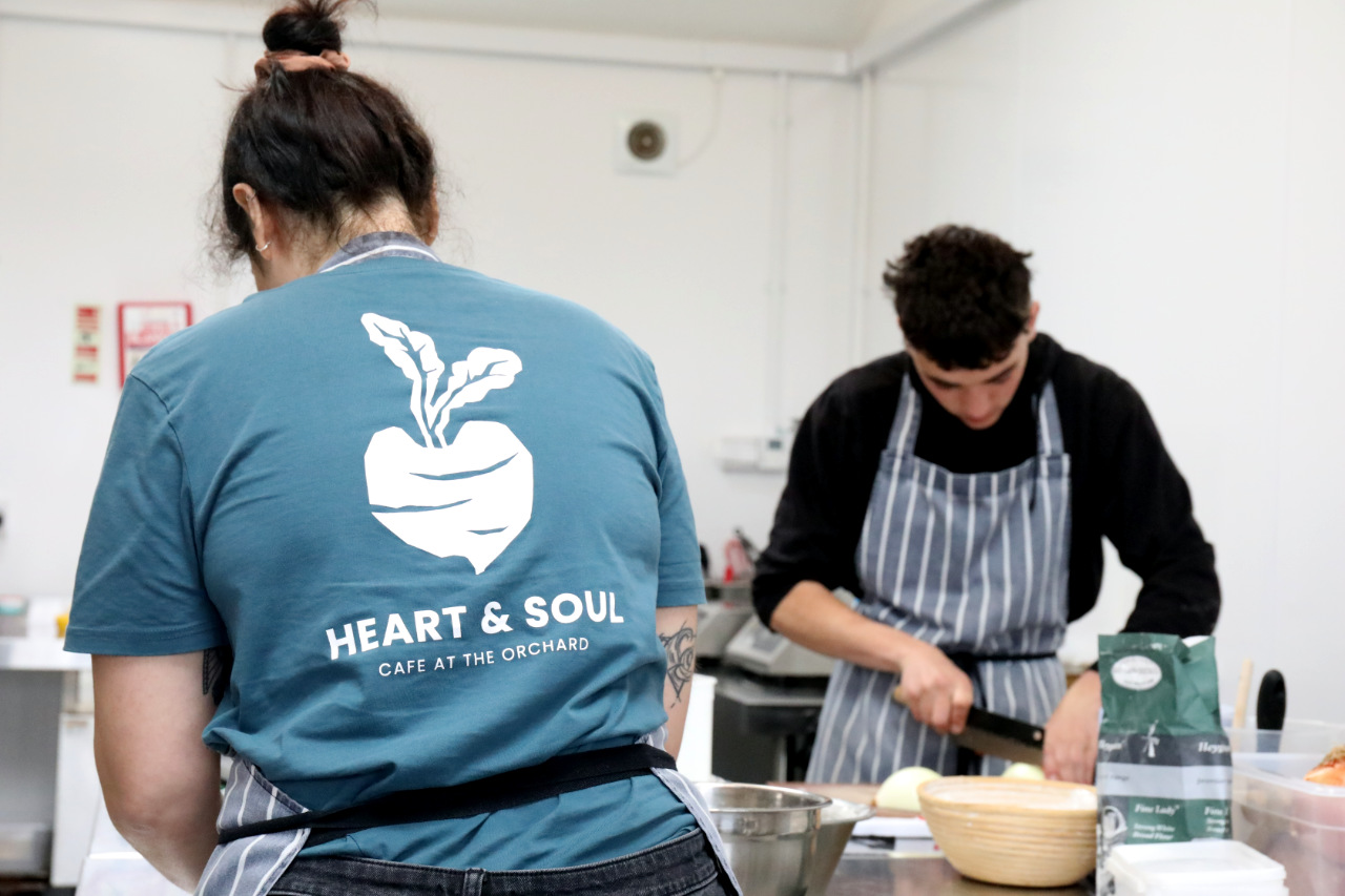 A person seen from behind wearing a "Heart & Soul Cafe at the Orchard" t-shirt, with a chef in a striped apron chopping onions in the kitchen behind them.