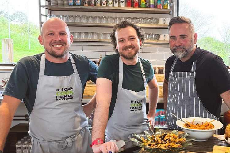 The Heart & Soul team smiling in the kitchen, two wearing aprons that read "If I can cook I can do anything," with prepared dishes on the counter in front of them.