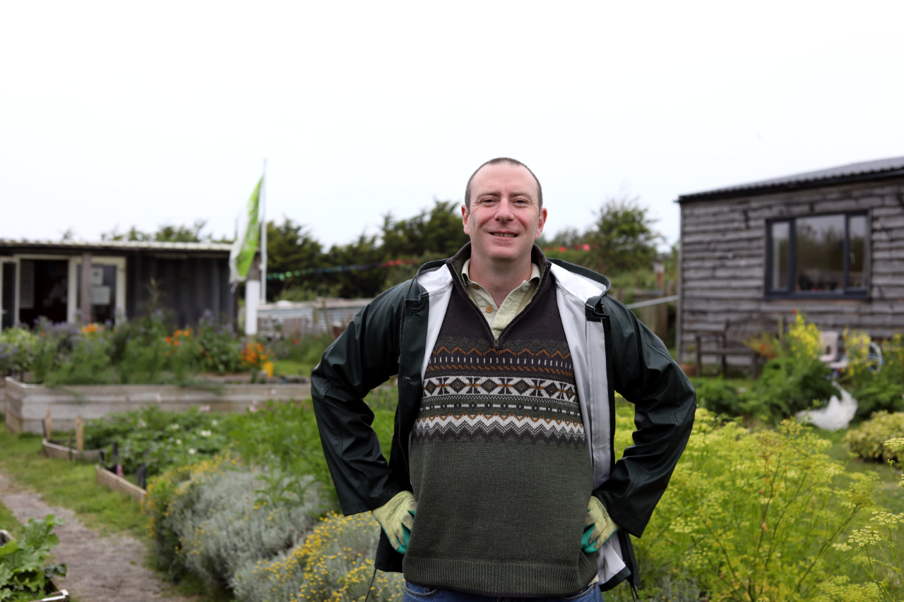 Matt, wearing a patterned jumper, stands with hands on hips smiling in an outdoor garden, with raised vegetable beds, herbs, and a wooden shed in the background.