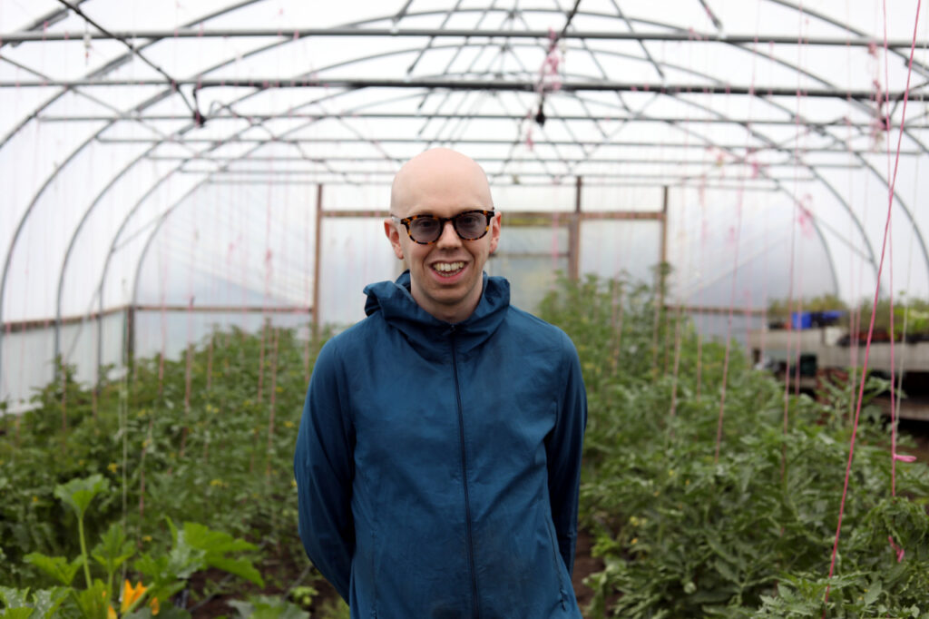 Ollie, wearing glasses and a blue hooded rain jacket, smiles at the camera while standing inside a polytunnel greenhouse, surrounded by rows of plants supported by string trellises.