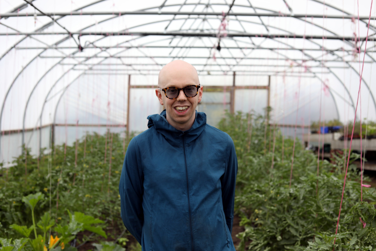 Ollie, wearing glasses and a blue hooded rain jacket, smiles at the camera while standing inside a polytunnel greenhouse, surrounded by rows of plants supported by string trellises.